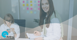 Two women in an office, one is smiling at the camera and the other is looking at something in her hands